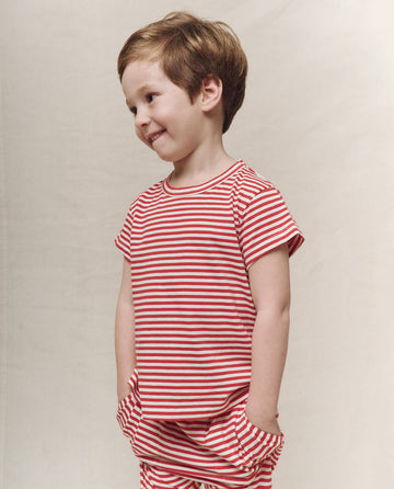 A young child smiles in a Sun Cherry Stripe Little Boxy Crew shirt and pants by THE GREAT. SP25 POLO LOUNGE LITTLE, set against a plain backdrop.