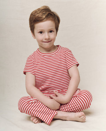 A child with short hair sits cross-legged in a Sun Cherry Stripe, exuding the relaxed style of The Little Boxy Crew by THE GREAT. SP25 POLO LOUNGE LITTLE, against a plain background.