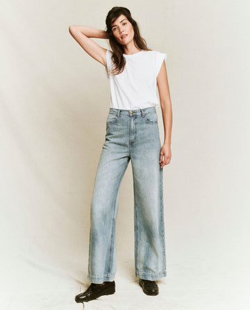 A person is standing against a plain background, wearing The Peak Shoulder Tee in True White from THE GREAT. FALL 22 KNITS collection; paired with light blue high-waisted wide-leg jeans and black shoes, with one hand resting on their head.