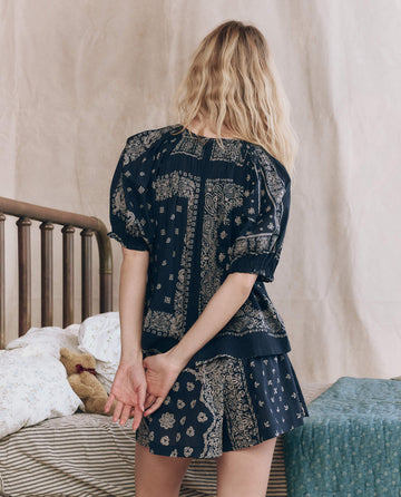 A woman with long, wavy blonde hair stands with her back to the camera, wearing THE GREAT. F25 SLEEP's Smocked Sleep Short in Bandana Patchwork next to a bed with floral bedding and a stuffed animal.