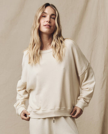 A person with long blonde hair is wearing The Teammate Sweatshirt in Washed White, designed for a relaxed fit by THE GREAT. FALL21 KNITS, and matching pants, standing in front of a beige backdrop.