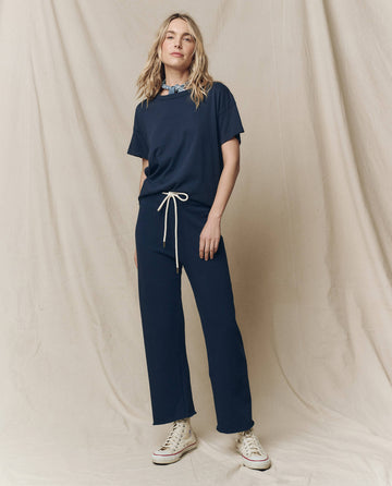 Against a beige backdrop, a person wears THE GREAT.'s Boxy Crew in True Navy with a drawstring waist, white high-top sneakers, and a neck scarf. Vintage tees underneath complete the effortlessly timeless vibe.