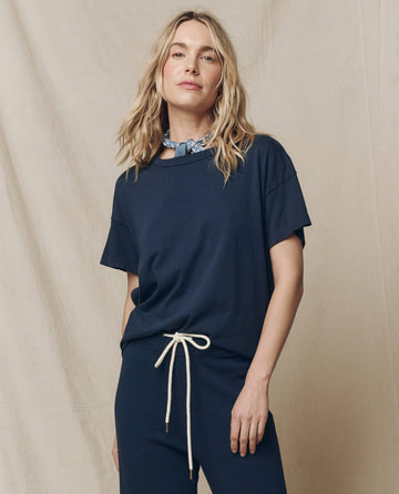 A person in The Boxy Crew Solid--True Navy by THE GREAT. FALL 23 KNITS, featuring a white drawstring and a light blue handcrafted scarf, stands against a beige backdrop.