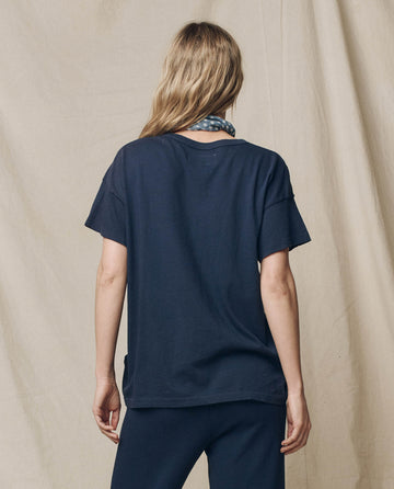 Facing away, a person with long hair wears THE GREAT.'s FALL 23 KNITS True Navy Boxy Crew cotton t-shirt and pants, accessorized with a handcrafted patterned scarf around the neck. Beige fabric backdrop.