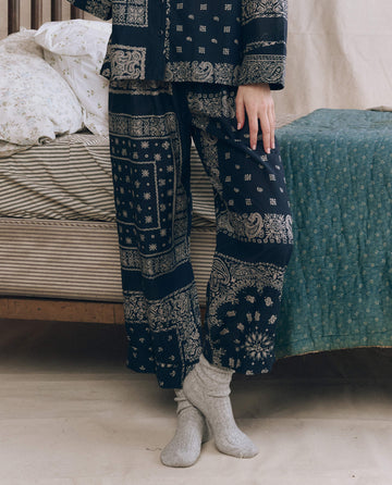 A person stands indoors by a bed with striped sheets and a green blanket, wearing THE GREAT. F25 SLEEP’s The Easy Sleep Pant in Bandana Patchwork print and gray socks.