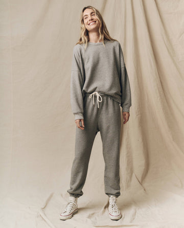A person wearing THE GREAT. FALL21 KNITS' "The Teammate Sweatshirt" in Varsity Grey, paired with matching pants and white sneakers, stands smiling against a beige fabric backdrop.