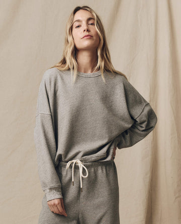 A person with long hair wearing The Teammate Sweatshirt in Varsity Grey stands against a neutral, textured backdrop.