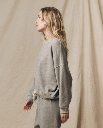 A person with long, wavy hair is standing in profile wearing The Teammate Sweatshirt in Varsity Grey and matching sweatpants from THE GREAT. FALL21 KNITS, handcrafted in Los Angeles, against a beige backdrop.