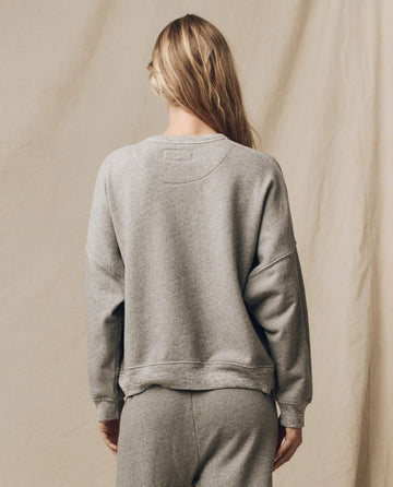A person with long hair, dressed in THE GREAT. FALL21 KNITS' The Teammate Sweatshirt in Varsity Grey and matching sweatpants, handcrafted in Los Angeles, is standing and facing away against a beige backdrop.
