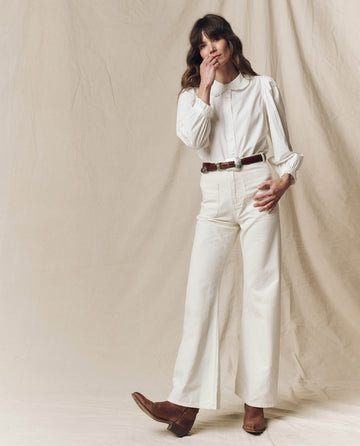 Wearing a white Hemingway Top by THE GREAT. PS25 with flared pants, complemented by a brown belt and shoes, a person stands elegantly against a beige backdrop, lightly touching their face.