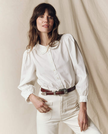 A person with long hair models The Hemingway Top by THE GREAT. PS25, paired with stylish pants against a neutral background, hand resting on hip.