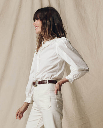 A person with long hair stands sideways, elegantly dressed in The Hemingway Top by THE GREAT. PS25, paired with chic pants and a brown belt, embodying timeless charm against a beige fabric backdrop.