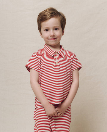A young child with short hair stands smiling with hands clasped in front, wearing THE GREAT. SP25 POLO LOUNGE LITTLE's "The Little Lacrosse Polo" in Sun Cherry Stripe, against a plain background.