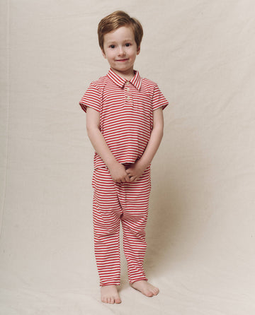 A child wears The Little Jersey Crop in Sun Cherry Stripe by THE GREAT. SP25 POLO LOUNGE LITTLE, standing barefoot on a neutral backdrop, smiling with hands clasped.