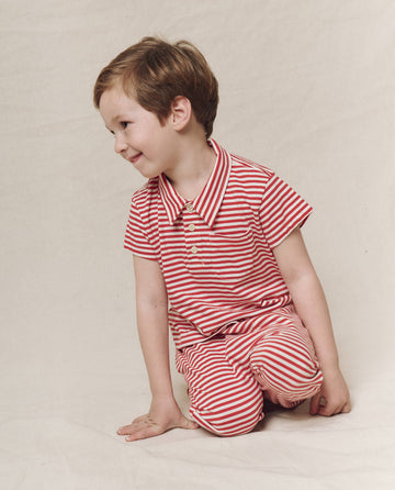 A child wearing The Little Lacrosse Polo in Sun Cherry Stripe by THE GREAT. SP25 POLO LOUNGE LITTLE kneels and smiles on a neutral background.