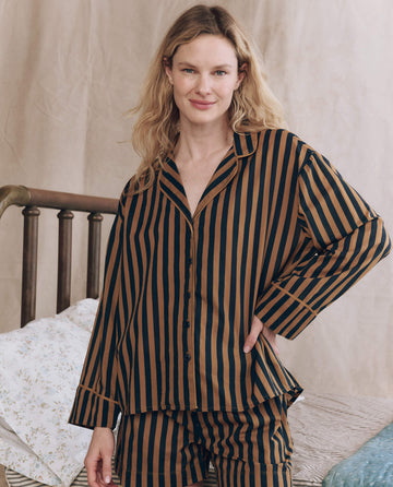 A woman stands indoors with her hand on her hip, wearing THE GREAT. F25 SLEEP The Pajama Shirt in Club Stripe—a brown and black vertically striped cotton sleepwear set—by a bed with patterned bedding.