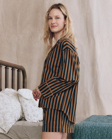 A woman stands by a bed wearing THE GREAT. F25 SLEEP’s The Sleep Boxer Short in Club Stripe, paired with a matching striped long-sleeve top, featuring an elasticated waistband, against neutral bedding and background.