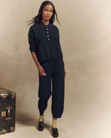 A person wears a dark long-sleeve collared top and The Lantern Pant in Dark Navy by THE GREAT. SP25 HIGH TWIST YARN. With hands in pockets, they sport light socks with dark shoes. A vintage trunk stands nearby against a neutral backdrop, completing this comfortable look.