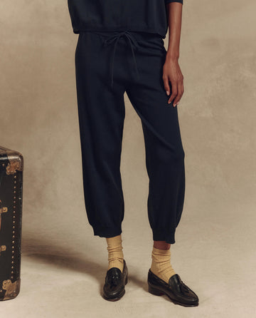 A person in The Lantern Pant by THE GREAT. SP25 HIGH TWIST YARN—Dark Navy, and black loafers stands beside a vintage trunk against a plain backdrop, effortlessly blending stylish pants with timeless charm.