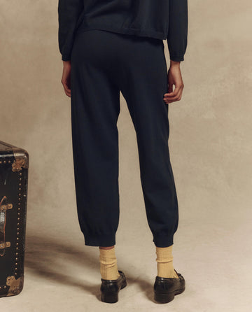 Clad in a cozy sweater and stylish THE GREAT. SP25 HIGH TWIST YARN’s The Lantern Pant in Dark Navy, accented by yellow socks and black shoes, a person stands near a vintage trunk, facing away from the camera.