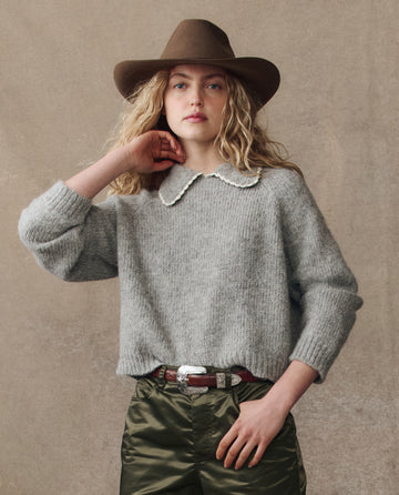 A person with long, wavy blonde hair wears THE GREAT. H25 COLLECTION's The Crochet Collar Pullover in Heather Grey, paired with a brown wide-brimmed hat and green belted pants, standing against a neutral background.