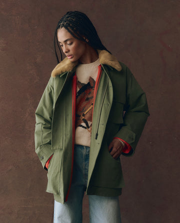 Wearing THE GREAT. F25 D1’s The Quilted Liner Parka - Army with Hot Orange, a patterned sweater, and light blue jeans, a person stands against a plain brown background, looking down with hands in jacket pockets.