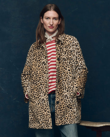 A woman stands against a dark background wearing THE GREAT. F25 COLLECTORS' The Car Coat. -- Vintage Leopard, a midcentury-inspired faux fur leopard print coat with quilted satin-lined pockets, over a red-and-white striped shirt and blue jeans.