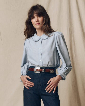 A person with long hair wearing The Hemingway Top in Light Chambray by THE GREAT. PS25 and dark jeans is standing in front of a beige backdrop.