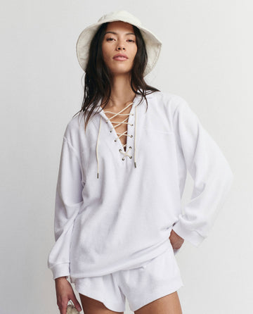 A person wears THE GREAT. SU25 MICRO TERRY’s Micro Terry Lace Up Tunic in True White, styled with a white bucket hat and shorts, posing against a plain background with one hand on their hip.