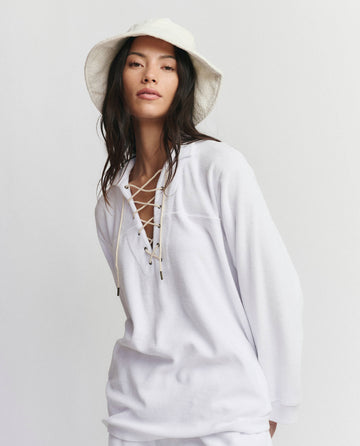 A woman wearing THE GREAT. SU25 MICRO TERRY The Micro Terry Lace Up Tunic in True White and a white bucket hat stands against a plain light background.