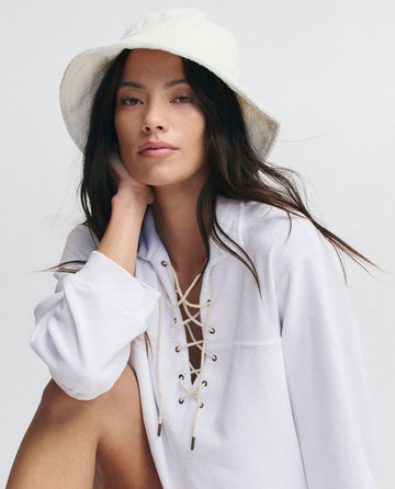 A woman with long brown hair wears THE GREAT. SU25 MICRO TERRY's The Micro Terry Lace Up Tunic in True White and rests her chin on her hand against a plain background, paired with a white bucket hat.
