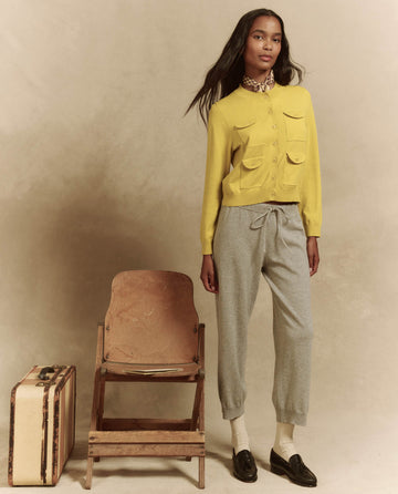 A person stands beside a wooden chair and suitcase, wearing a yellow cardigan and fashionable THE GREAT. SP25 HIGH TWIST YARN's The Lantern Pant in Heather Grey, paired with black shoes against a neutral backdrop.
