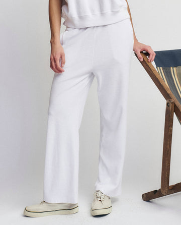 Person wearing THE GREAT. SU25 MICRO TERRY The Micro Terry Victory Sweatpant in True White and white sneakers, standing next to a wooden and fabric chair against a plain background.