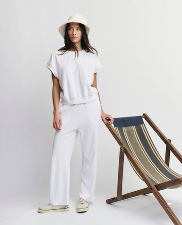 A woman wearing THE GREAT. SU25 MICRO TERRY The Micro Terry Wedge Sweatshirt in True White stands beside a wooden deck chair with striped fabric, set against a plain white background.