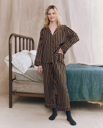 A woman stands by an unmade bed, wearing THE GREAT. F25 SLEEP's The Easy Sleep Pant in Club Stripe—loose-fitting, high-waist black and brown striped sleepwear—against a neutral fabric backdrop.
