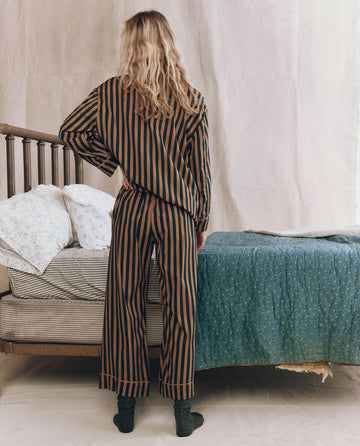 A person with long blond hair stands facing away, wearing THE GREAT. F25 SLEEP The Easy Sleep Pant in Club Stripe next to a bed with patterned bedding in a softly lit room.