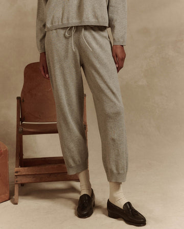 A person in The Lantern Pant in Heather Grey by THE GREAT. SP25 HIGH TWIST YARN, paired with a gray sweatshirt, brown loafers, and white socks, stands next to a wooden folding chair against a soft neutral background.