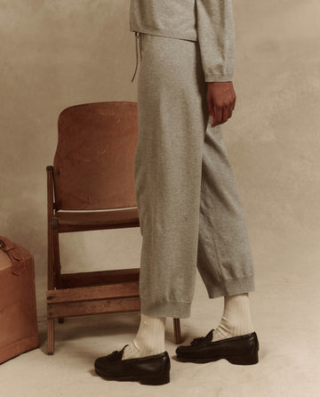 A person in The Lantern Pant - Heather Grey from THE GREAT., paired with black shoes, stands beside a wooden chair and suitcase against a beige background, exuding effortless fashion.