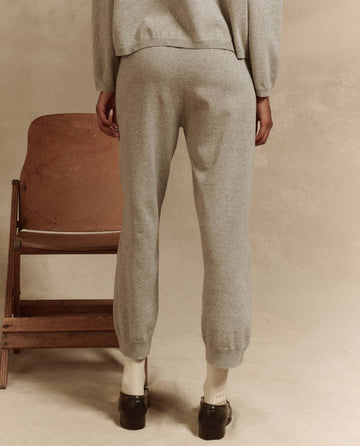 A person stands with their back turned, wearing The Lantern Pant in heather grey by THE GREAT. SP25 HIGH TWIST YARN. Complementing the look are black shoes and white socks, with a wooden chair to their right completing the contemporary ensemble.