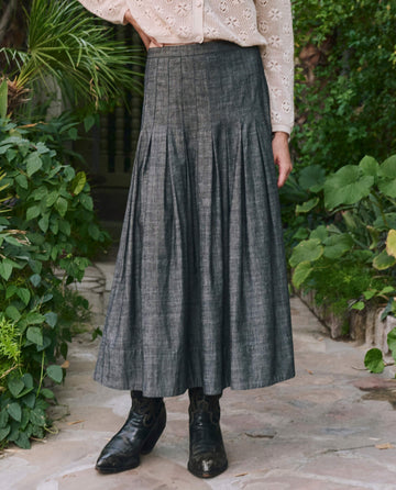 A person wears a light long-sleeve blouse, black boots, and THE GREAT. PF25 The Rhythm Skirt in Smoke Wash, standing on a stone path surrounded by green plants.