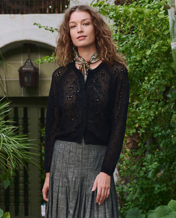 A woman with curly hair stands in a garden surrounded by greenery, wearing THE GREAT. PF25's The Crochet Cardigan in black, paired with a patterned scarf and gray pleated skirt.