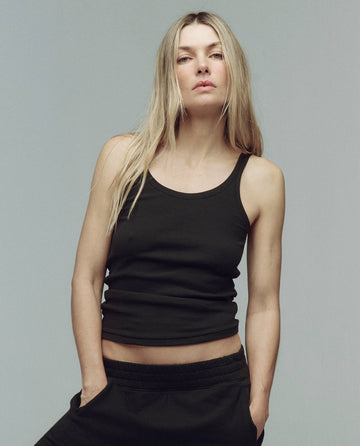 A person with long blonde hair wears THE GREAT. SU25 BABY RIB’s The Baby Rib Tank. -- Almost Black and black pants, standing against a plain gray background with hands in pockets.