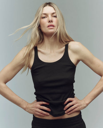 A person with long blonde hair stands with hands on hips against a neutral background wearing THE GREAT. SU25 BABY RIB "The Baby Rib Tank." in Almost Black.
