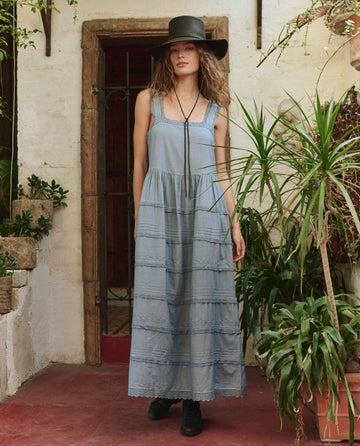 A woman in THE GREAT. PF25's The Reverie Dress in Pale Blue, styled with a black wide-brim hat and black boots, stands indoors surrounded by green potted plants.
