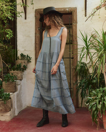 A person wearing THE GREAT. PF25 The Reverie Dress in Pale Blue, paired with a black hat and boots, stands on a red floor among potted green plants and a rustic doorway, making a chic fashion statement.