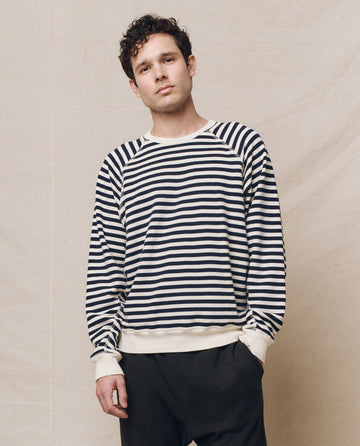Wearing THE GREAT. SP25 POLO LOUNGE MEN's The Men's College Sweatshirt in Navy Sailor Stripe, paired with dark pants, a person stands against a plain background.