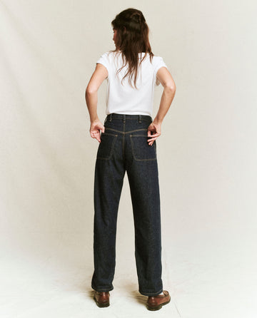 A person stands with their back to the camera, hands resting on the back pockets of their dark blue jeans. They’re wearing The Little Tee in True White from THE GREAT. SP22 APRILCAP that pairs perfectly as a layering piece, complemented by brown shoes.