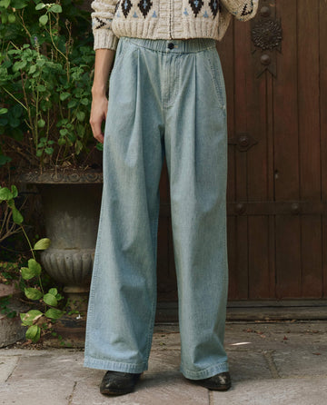 A person wearing THE GREAT. PF25 The Sawyer Pant in Boiler Wash, a patterned sweater, and black shoes stands on a stone path in front of a wooden door with plants nearby.
