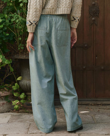 A person stands outdoors facing away, wearing a textured beige sweater and THE GREAT. PF25 The Sawyer Pant in Boiler Wash—showcasing an effortless, relaxed style against a backdrop of a wooden door and greenery.