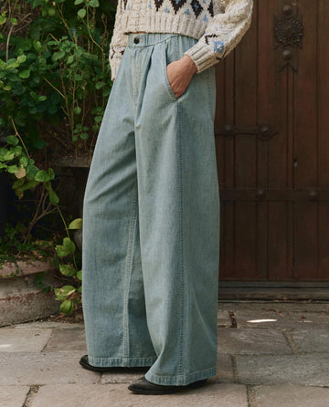 A person wearing a patterned sweater and The Sawyer Pant in Boiler Wash by THE GREAT. PF25 stands on a stone patio with hands in pockets, framed by green plants and a wooden door, showcasing their relaxed style.